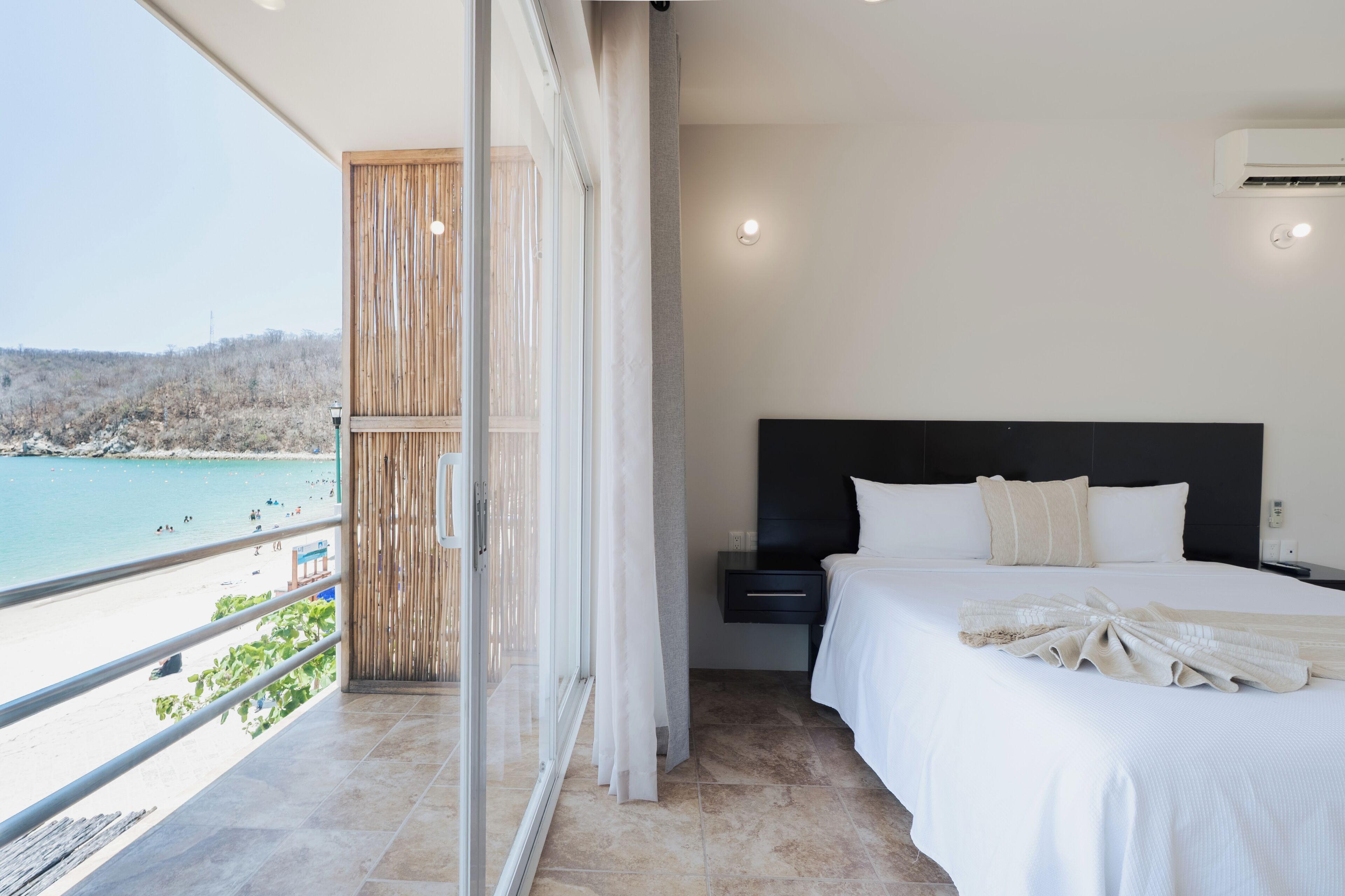 A bedroom with a bed, a nightstand, a view of a beach, and a balcony with a railing.