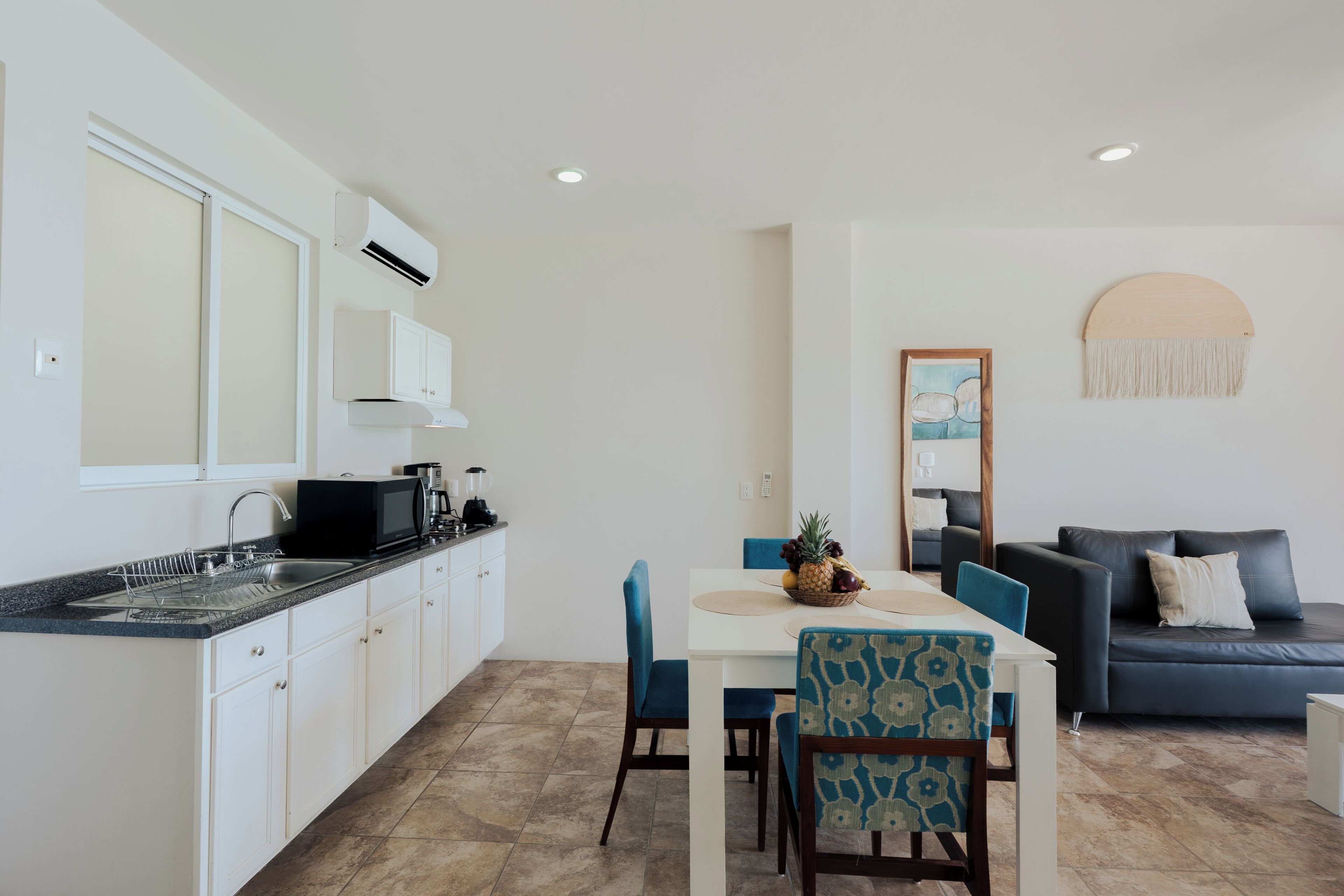 Una cocina moderna con armarios blancos, un microondas negro y un fregadero. Hay una zona de comedor con una mesa blanca, sillas azules y un cuenco de frutas. En la sala de estar se encuentra un sofá oscuro y un espejo redondo.