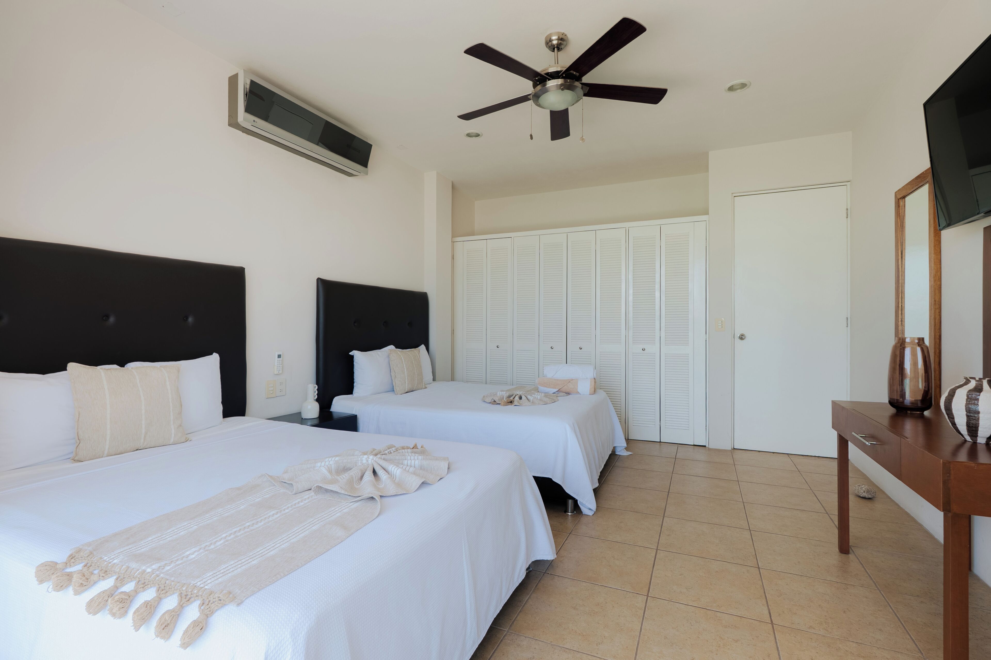 Habitación de hotel con dos camas, ventilador de techo, televisor, armario y mesita de noche.
