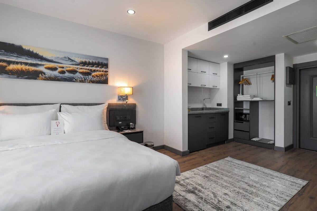 Habitació d'hotel moderna amb un llit gran, una tauleta de nit, un quadre, una petita cuina i un armari gris.