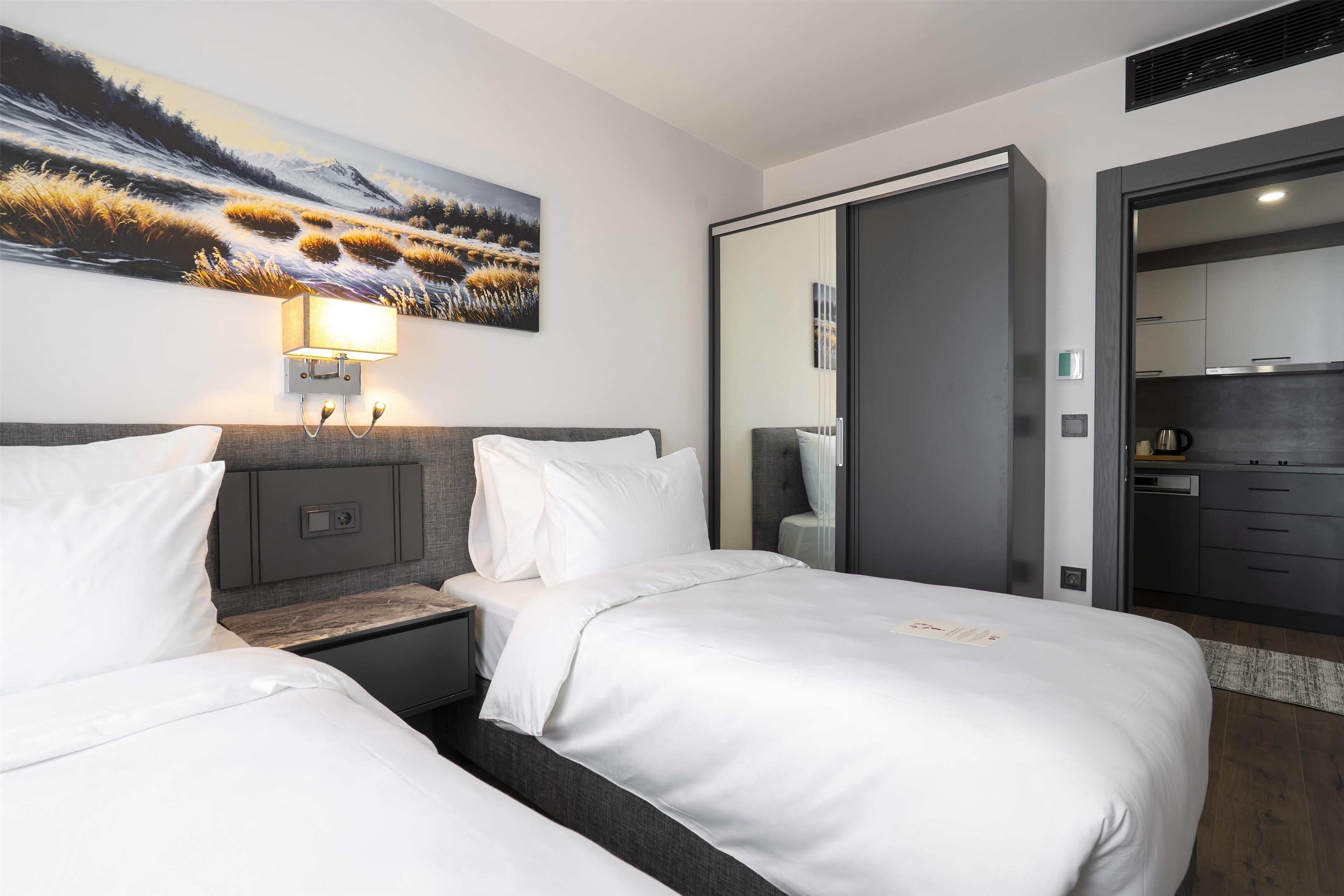 A hotel room with two beds, a nightstand, a wall-mounted lamp, a painting, and a view of the kitchen through a door.