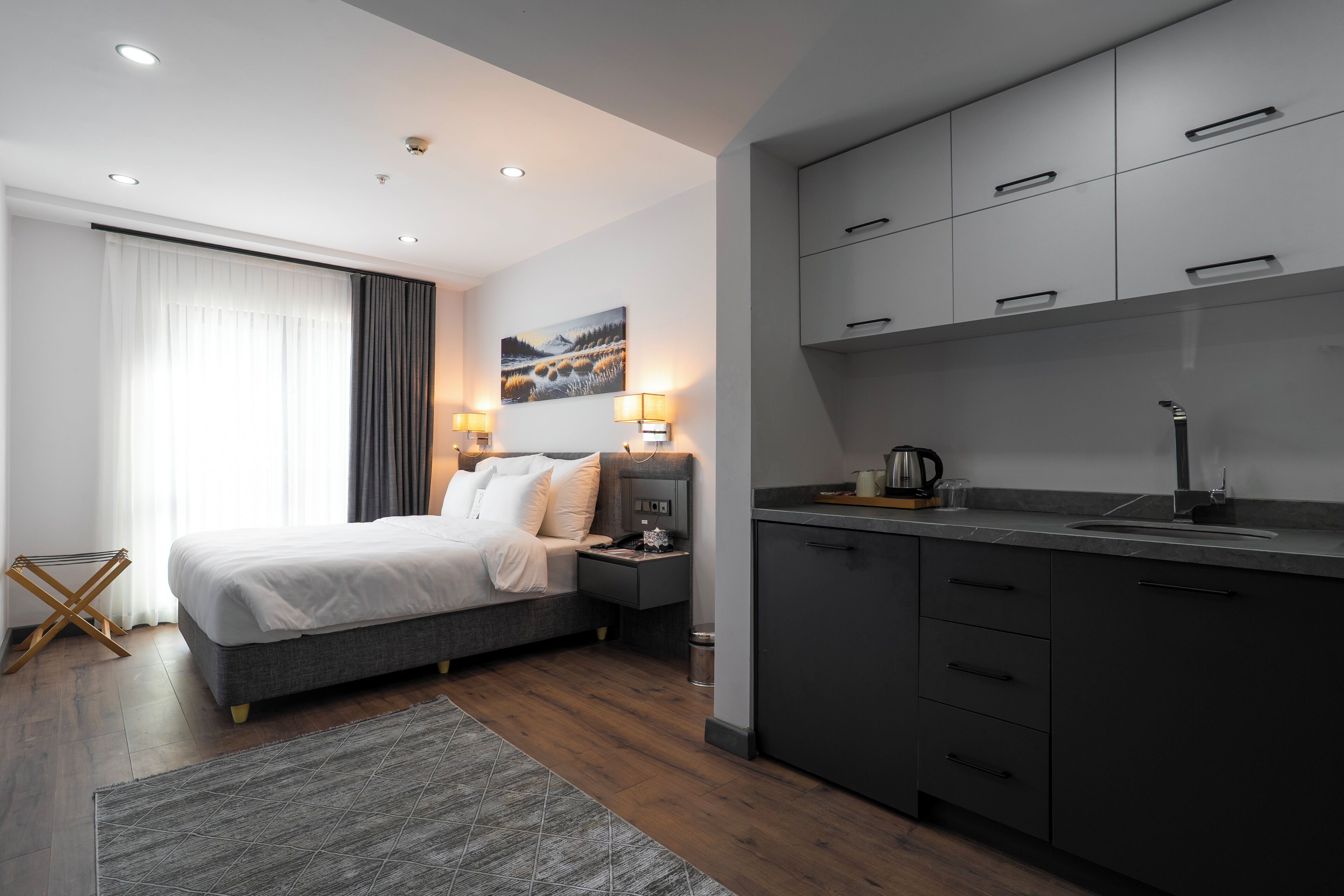 A modern hotel room with a large bed, a small desk, a kitchenette, and a painting on the wall.