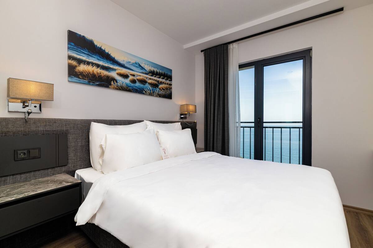 Habitació d'hotel moderna amb un llit gran, dues taules de nit amb làmpades, un quadre a la paret i vista al mar a través d'una finestra ampla.