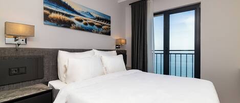 A modern hotel room with a large bed, two bedside tables with lamps, a painting on the wall, and a view of the sea through a large window.