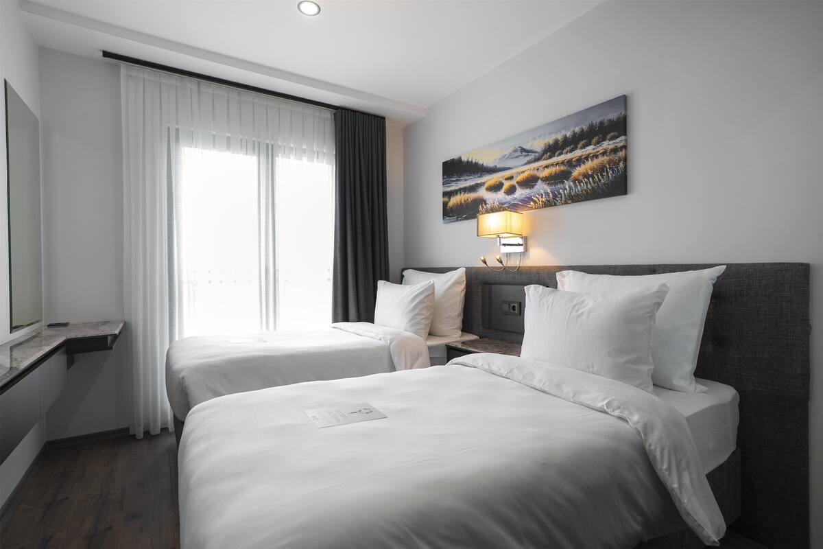 Habitació d'hotel amb dos llits, una finestra gran amb cortines, un quadre a la paret i una làmpada de nit.