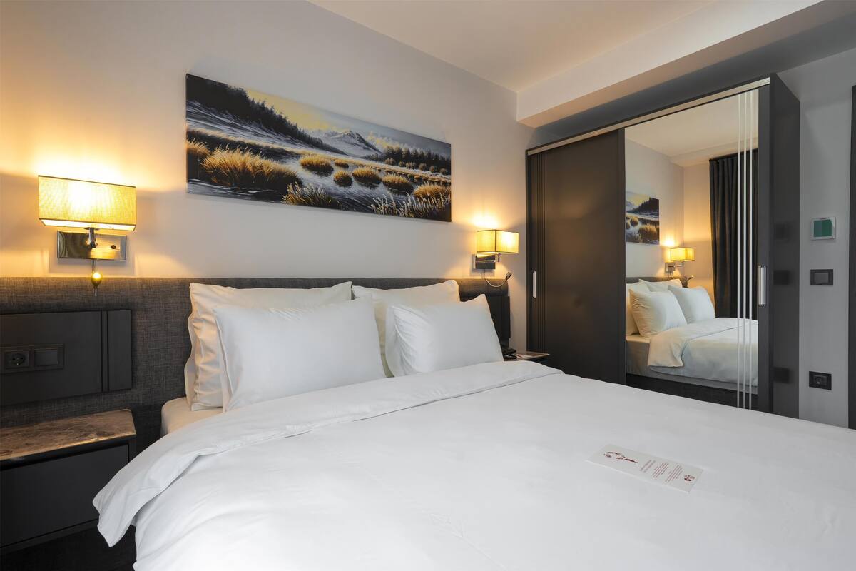Habitació d'hotel amb un llit gran, dues làmpades de nit, un quadre a la paret i un mirall que reflecteix l'habitació.