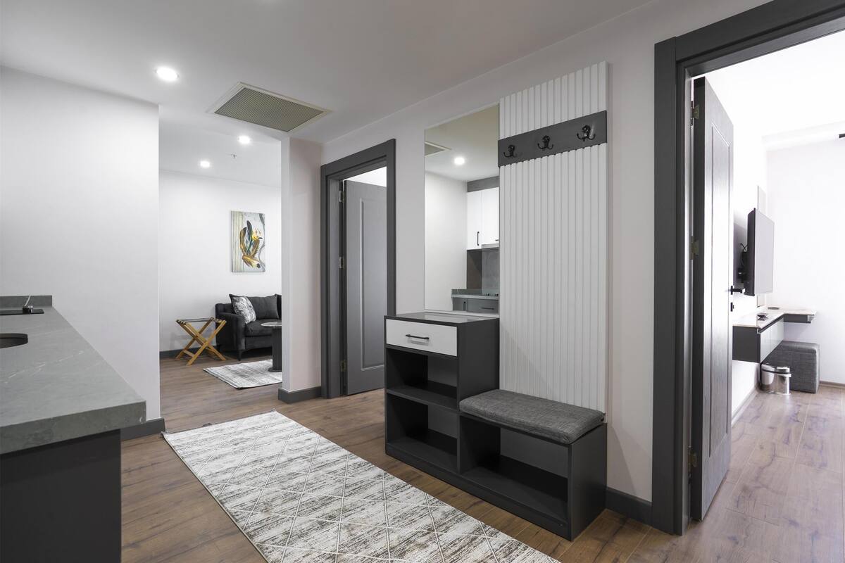 Habitació moderna i ben il·luminada amb un sofà gris, un escriptori petit amb una cadira, una banca i un mirall gran.