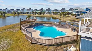 Ein Schwimmbecken mit Holzterrasse, umzäunt, mit Blick auf Häuser und ein Gewässer im Hintergrund.