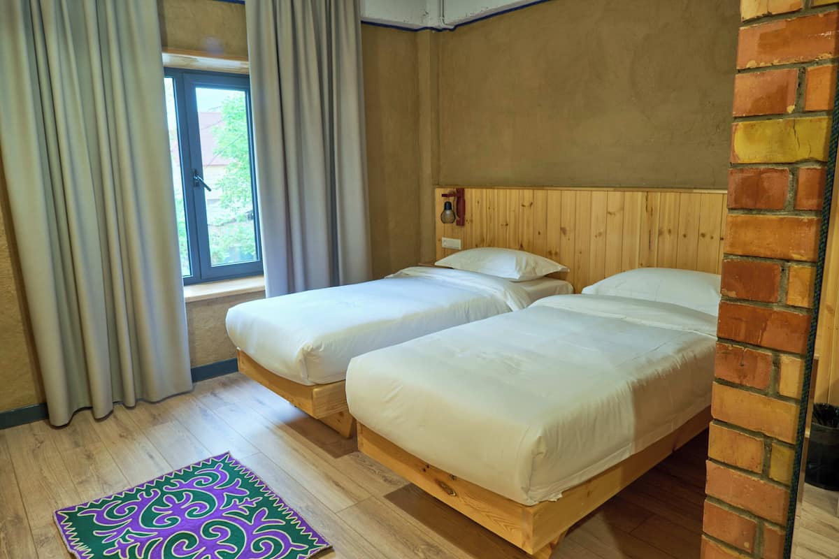 A room with two wooden beds, a window with curtains, a patterned rug, and a brick wall.
