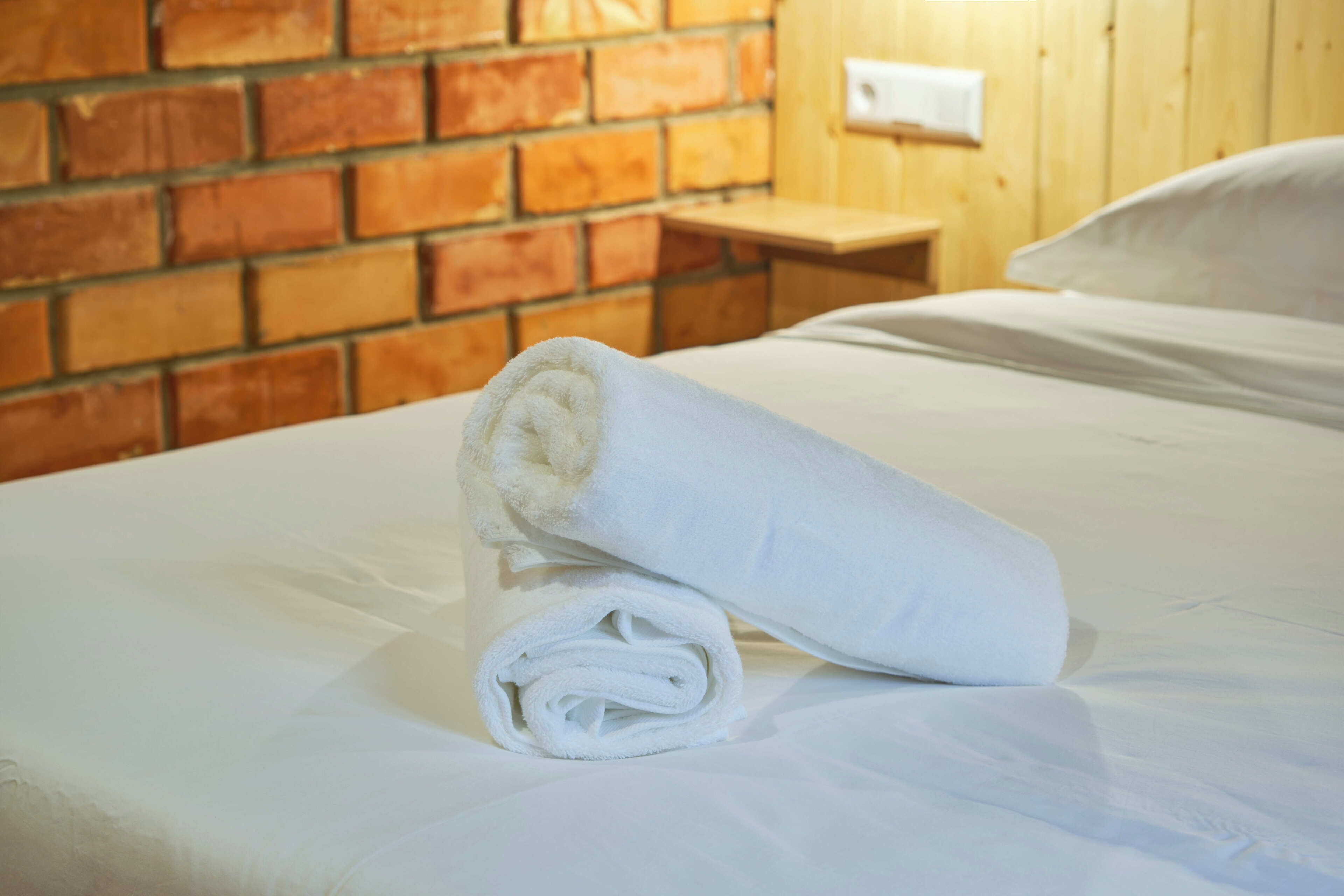 Una cama bien hecha con una toalla blanca enrollada y una mesita de noche de madera con un interruptor de luz.