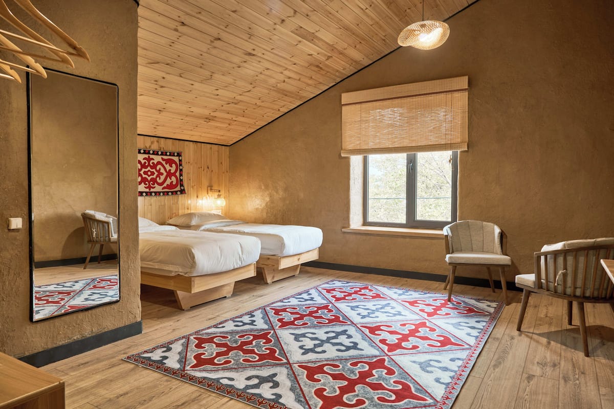 A bedroom with a wooden ceiling, a large mirror, two beds, a window with a bamboo blind, and a patterned rug.