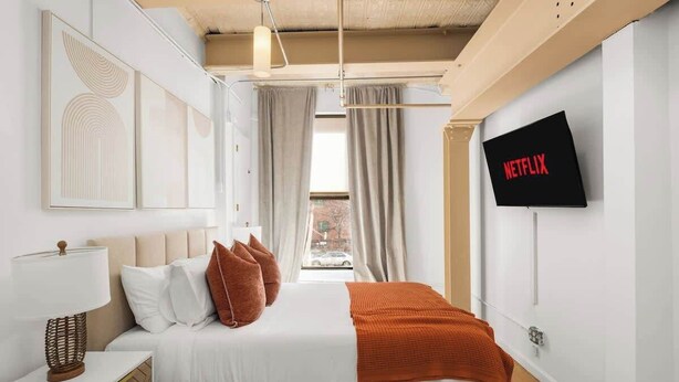 A modern bedroom with a bed, a bedside table, a lamp, a TV mounted on the wall, and a window with curtains. - Spacious 3 Bedroom Gem in Trendy Dumbo! (Brooklyn)