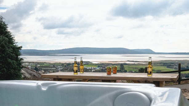A wooden table with bottles of Corona Extra and Liptun beer, overlooking a body of water and distant hills. - Raphael House (Burry Port)