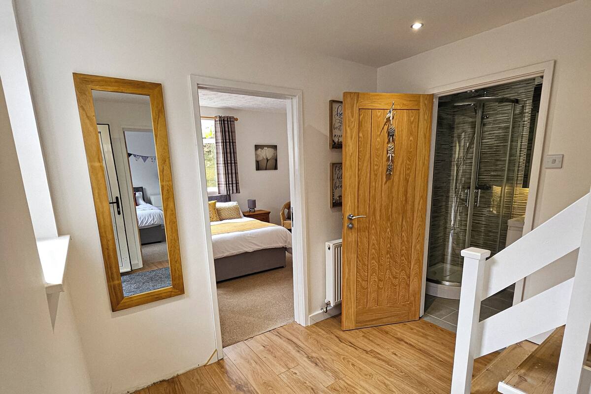 A hallway with wooden flooring, a mirror, a door leading to a bedroom, and a shower area.