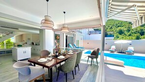 Un espace repas moderne avec vue sur la piscine, comprenant une table à manger pour six personnes, une cuisine en arrière-plan et un coin salon au bord de la piscine agrémenté de parasols.