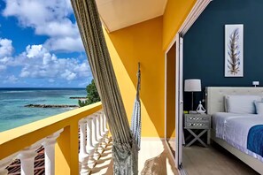 Een balkon met uitzicht op de oceaan, een hangmat, een bed met wit beddengoed, een nachtkastje en een muur met een verenkunstwerk.