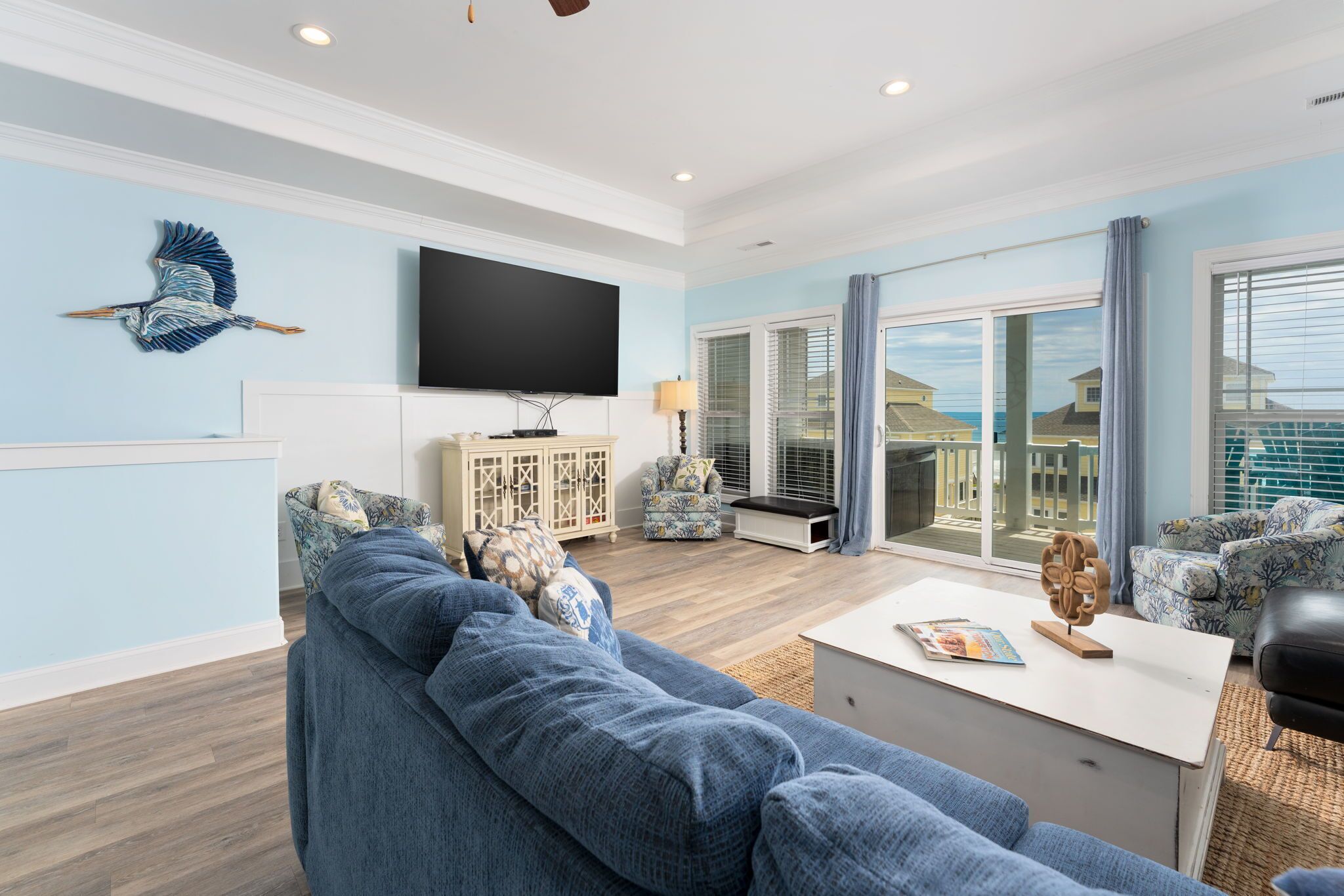 A spacious living room with a large sofa, a coffee table, and a television. There is a window with a view of the ocean and a balcony.