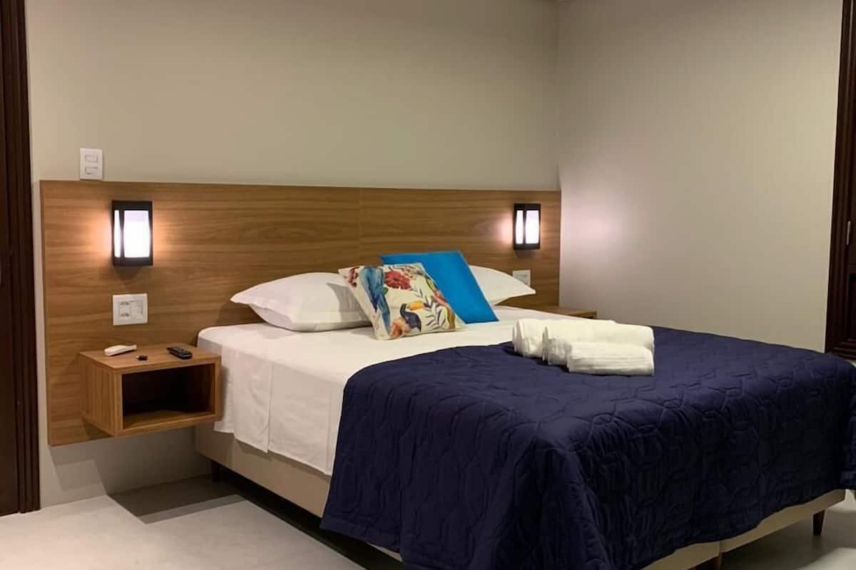 Una cama bien hecha con una colcha azul y almohadas, una mesita de noche con un control remoto y dos lámparas fijadas a la pared.