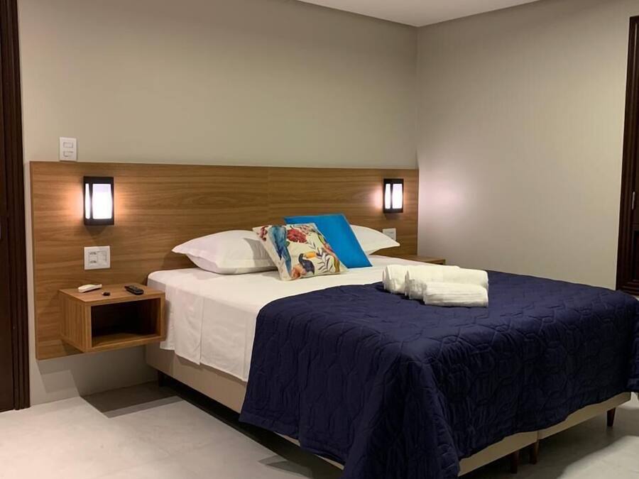 Una cama bien hecha con una colcha azul y almohadas, una mesita de noche con un control remoto y dos lámparas fijadas a la pared.