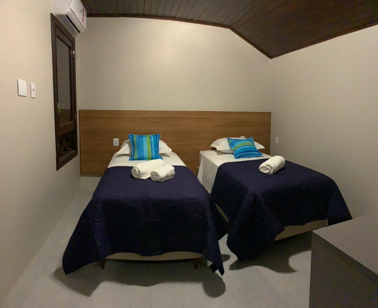 Una habitación pequeña y moderna con dos camas individuales, cada una con colcha y almohadas azules. Tiene un cabecero de madera, una ventana pequeña y un equipo de aire acondicionado instalado en la pared.