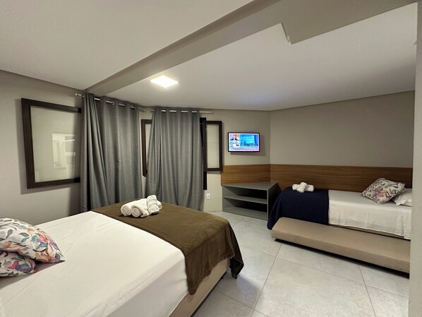 Habitación de hotel con una cama grande, una cama más pequeña, un banco, un televisor y una ventana con cortinas.