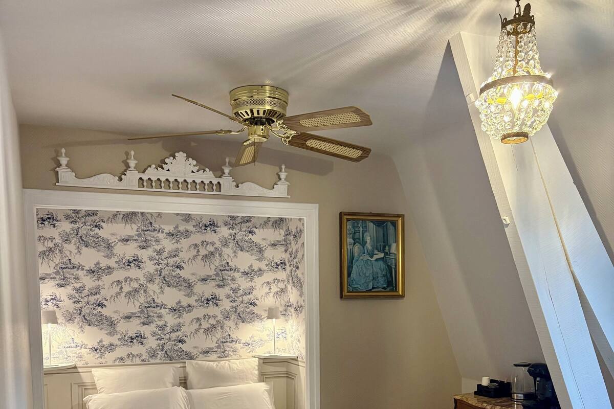 Een slaapkamer met een bed, een plafondventilator, een kroonluchter, een nachtkastje en een schilderij aan de muur.