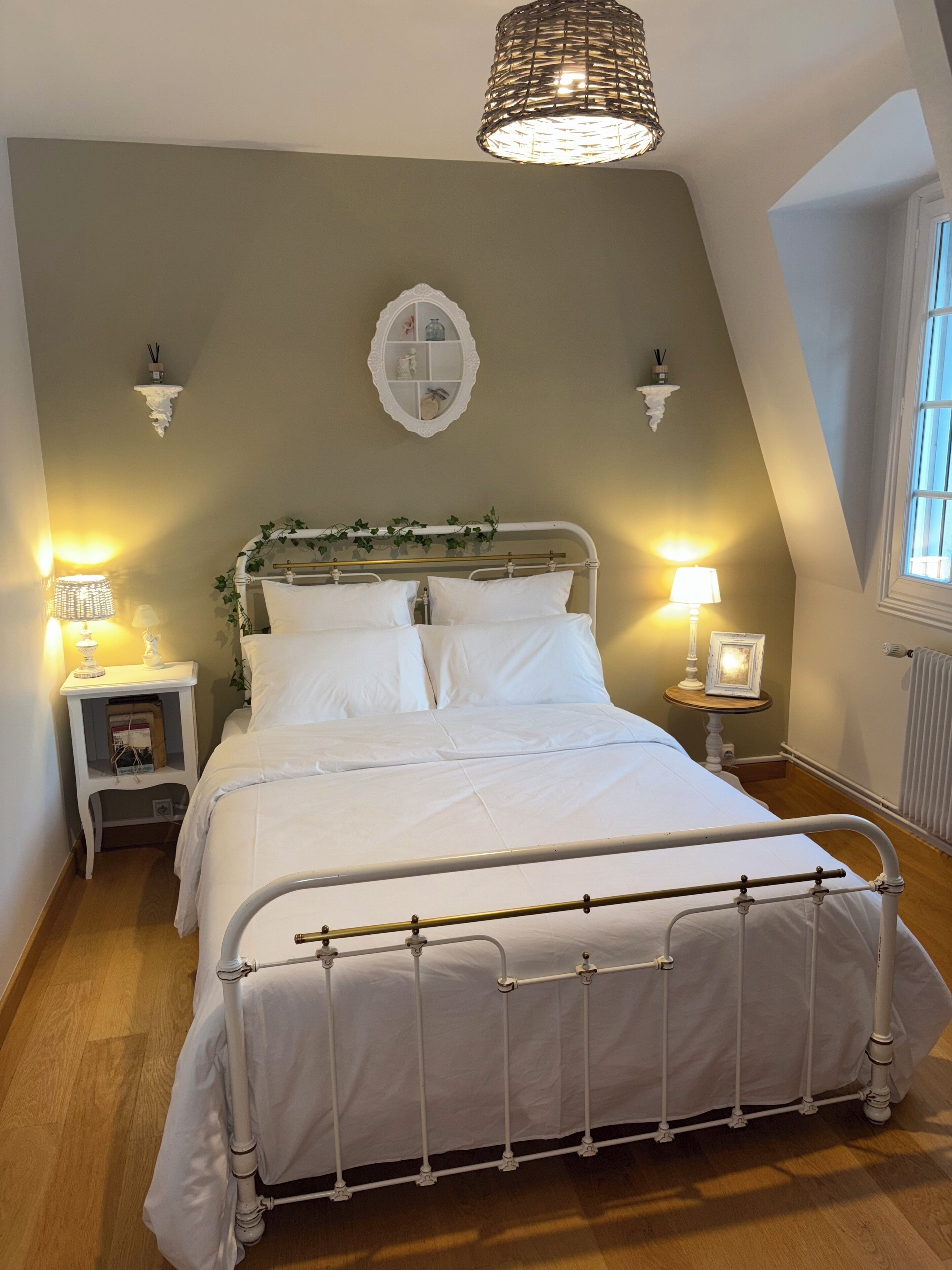 Ein Schlafzimmer mit einem Bettgestell aus weißem Metall, zwei Nachttischen mit Lampen, einem dekorativen Wandschrank und einem Fenster mit Jalousien.