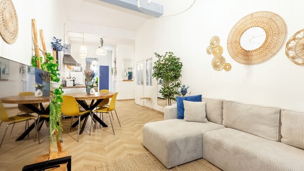 Ein modernes Wohnzimmer mit einem Ecksofa, einem Essbereich mit einem Holztisch und gelben Stühlen sowie dekorativen Wandbildern.
