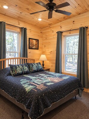 A wooden cabin room with a bed, a ceiling fan, a window with curtains, and a painting on the wall.