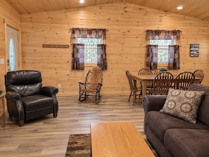 A log cabin interior with a leather chair, a rocking chair, a dining table with chairs, a sofa with a patterned pillow, and two windows with curtains.