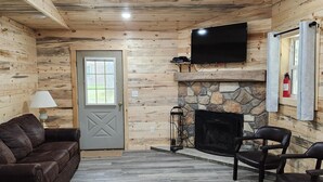 A log cabin interior with a stone fireplace, wooden walls, a mounted TV, and a cozy seating area.