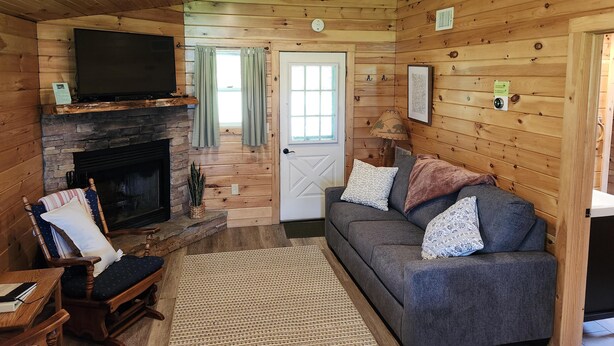 A cozy cabin interior with a stone fireplace, wooden walls, a gray sofa, and a television mounted on the wall. - Cozy Sugar Pine Cottage (Vowinckel)