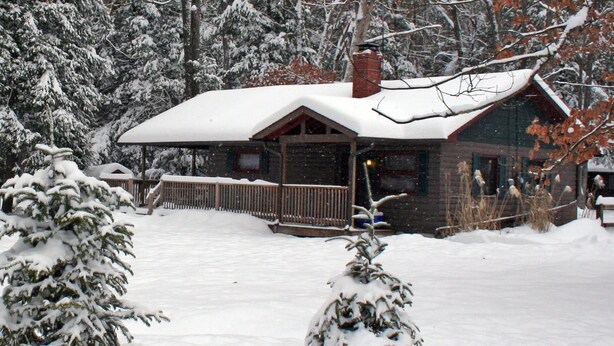 En stuga av trä med ett snötäckt tak och en veranda, omgiven av snötyngda träd.