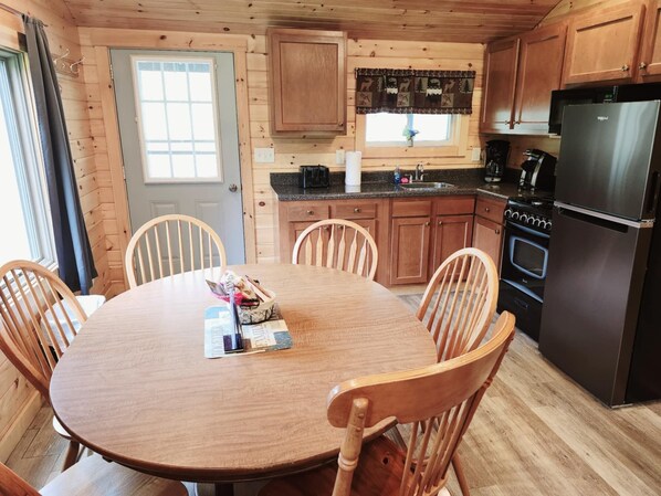 A wooden cabin interior with a round dining table, four chairs, a kitchen with a refrigerator, stove, and cabinets.