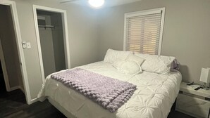 Un dormitorio con cama, ventilador de techo, ventana con persianas y mesita de noche con lámpara.
