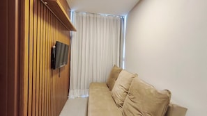 Ein Zimmer mit einer Holzwand, einem Wand-Fernseher, einem beigen Sofa und einem Fenster mit durchsichtigen Vorhängen.