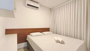 Quarto de hotel com cama, cabeceira de madeira, ar condicionado, janela com cortinas e duas toalhas enroladas sobre a cama.