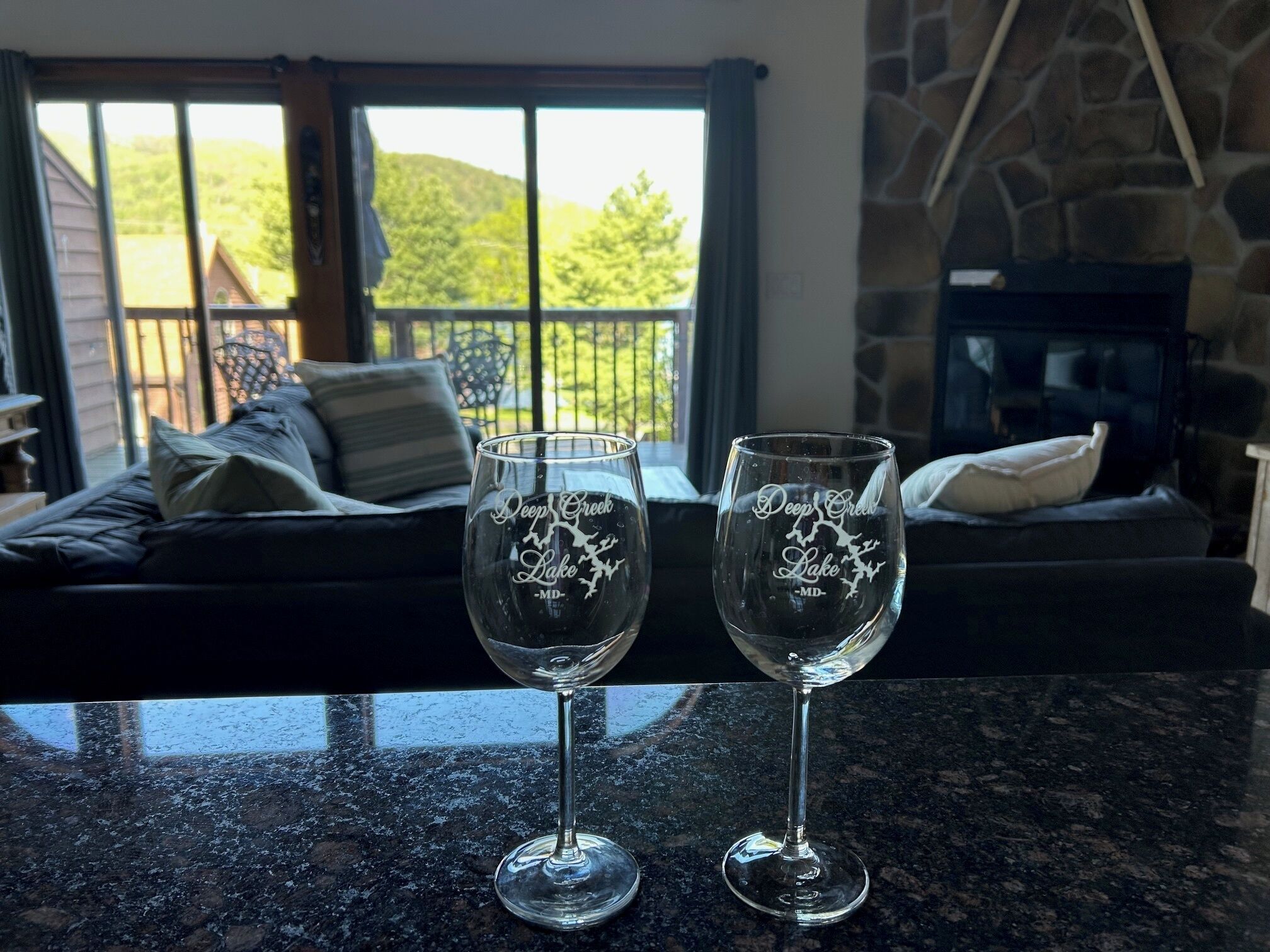 Zwei Weingläser mit der Gravur „Deep Creek Lake - MD“, platziert auf einer Granitarbeitsplatte in einem Raum mit Kamin und Blick auf einen Balkon mit einer Couch und Kissen.