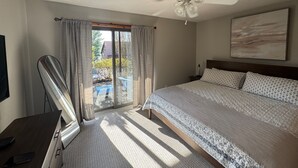 Un dormitorio con cama, ventilador de techo, ventana grande con cortinas y vistas al exterior.