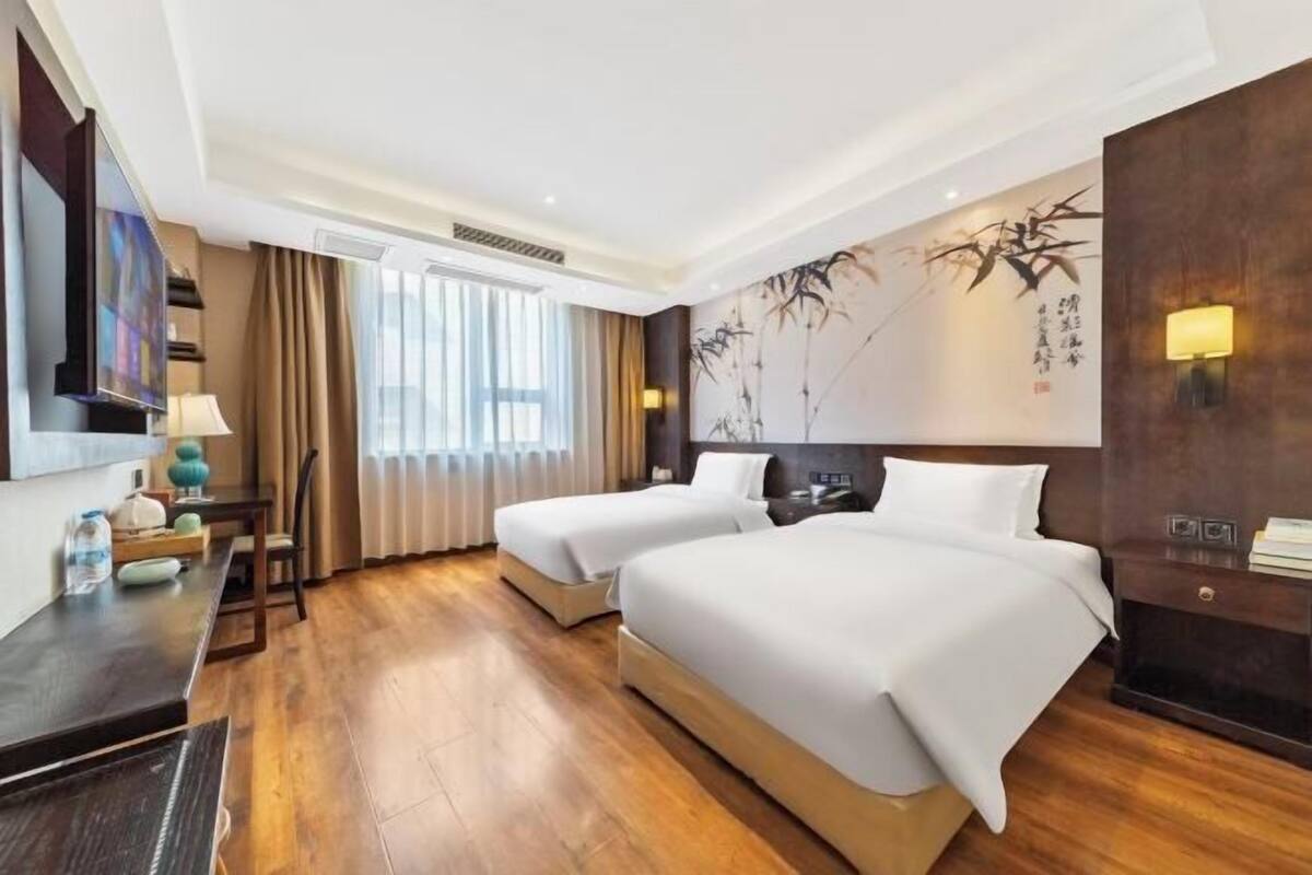 Habitació d'hotel amb dos llits, una escriptori, un televisor i una finestra gran amb cortines.