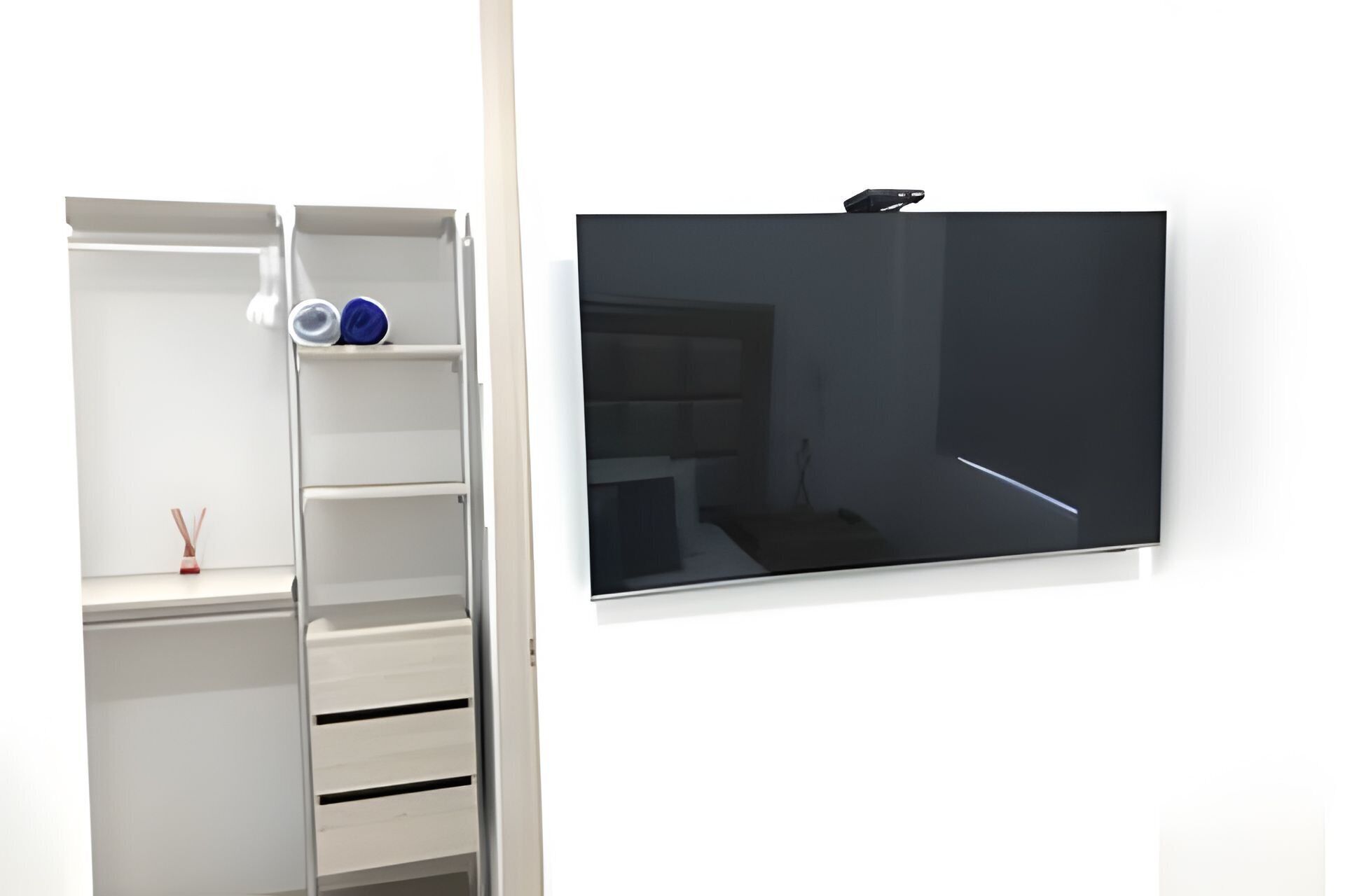Una habitación blanca con una televisión montada en la pared, un armario blanco con estantes y una mesita con un objeto decorativo.