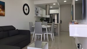 Una sala de estar moderna con un sofá negro, un comedor con sillas blancas, una cocina con electrodomésticos de acero inoxidable y un reloj en la pared.