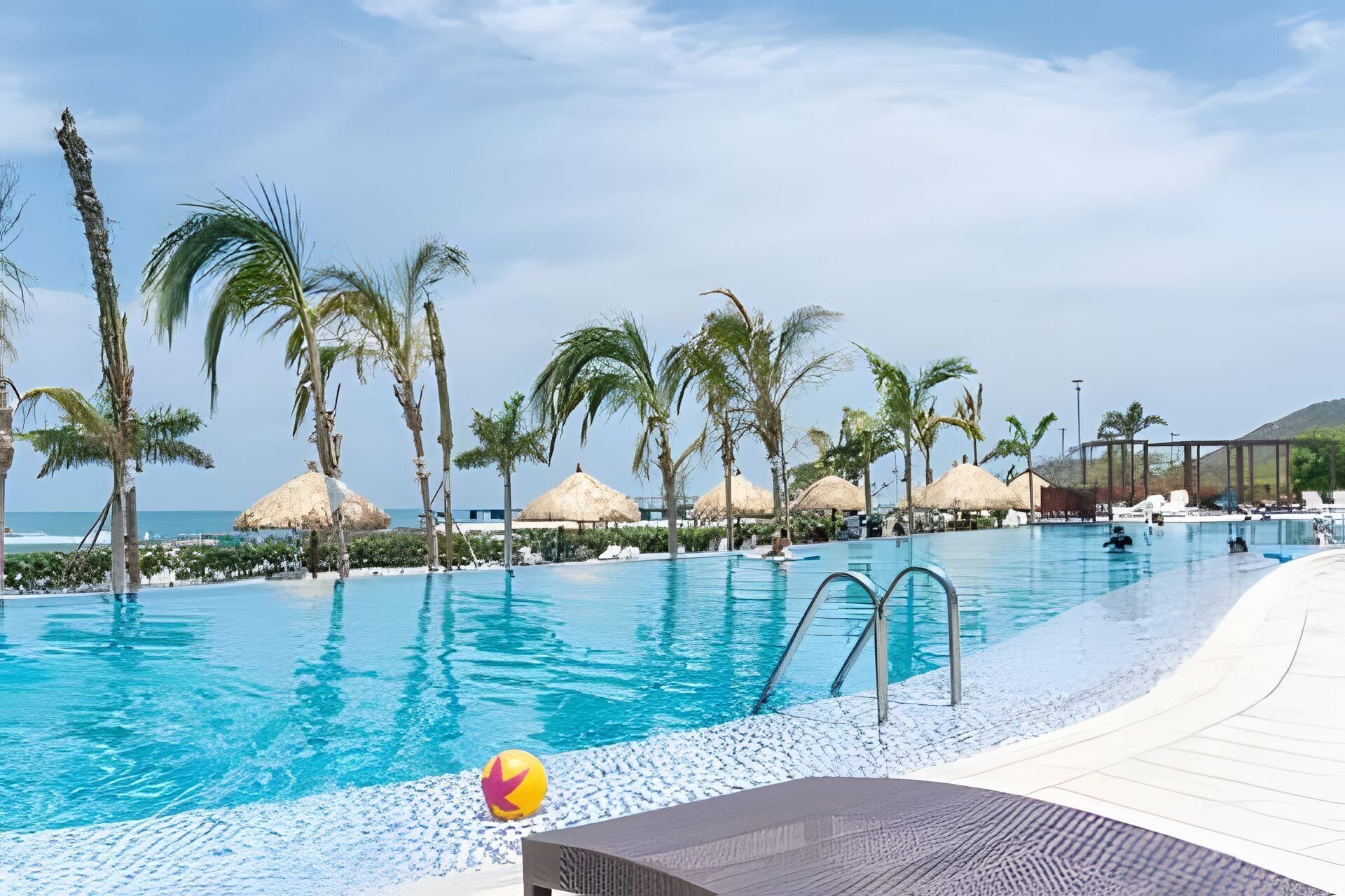 A poolside area with a clear view of the ocean, palm trees, and thatched umbrellas.