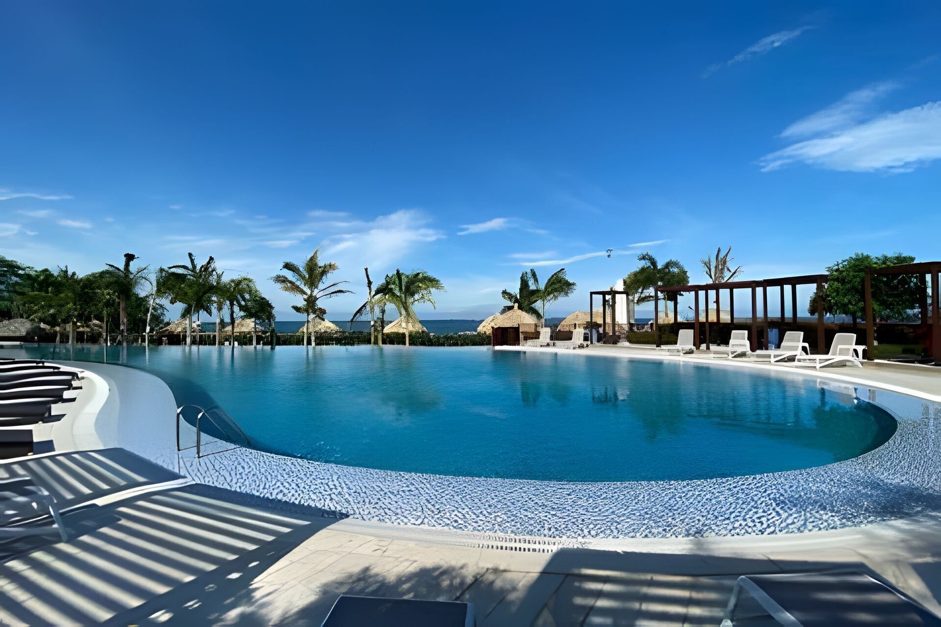 Une grande piscine aux eaux cristallines, bordée de chaises longues et de palmiers, et entourée d’une pergola en bois et de constructions à toits de chaume.