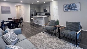 A modern living room with a gray sofa, a gray armchair, and a small rug. There is a ceiling fan, recessed lighting, and a kitchen in the background. - Bright Modern Retreat Near DIA & Gaylord Resort (Denver)