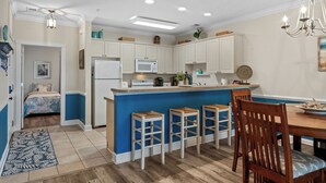 A modern kitchen with white cabinets, a blue accent wall, a central island with stools, and a dining area with a wooden table and chairs. - Bright and Modern 3BR Family Friendly Condo with pool, golf, Ripken Experience. (Myrtle Beach)