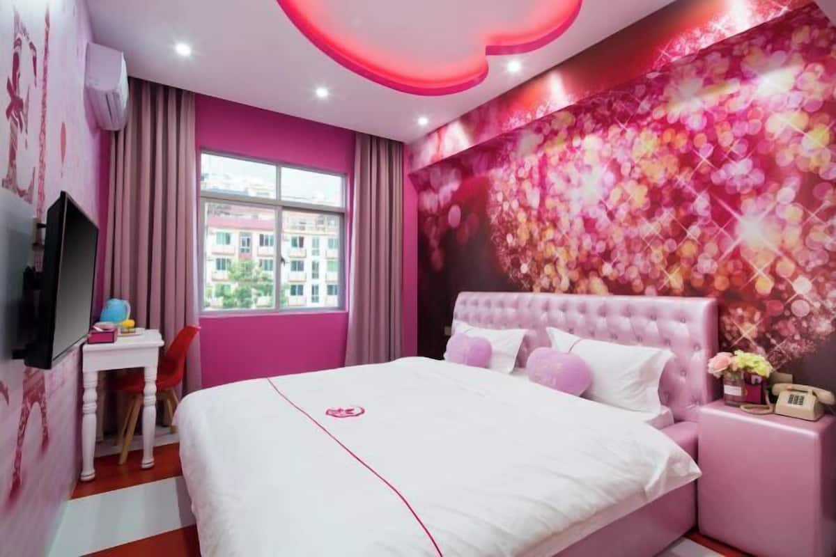 A pink-themed hotel room with a bed, a desk, a chair, and a large window.