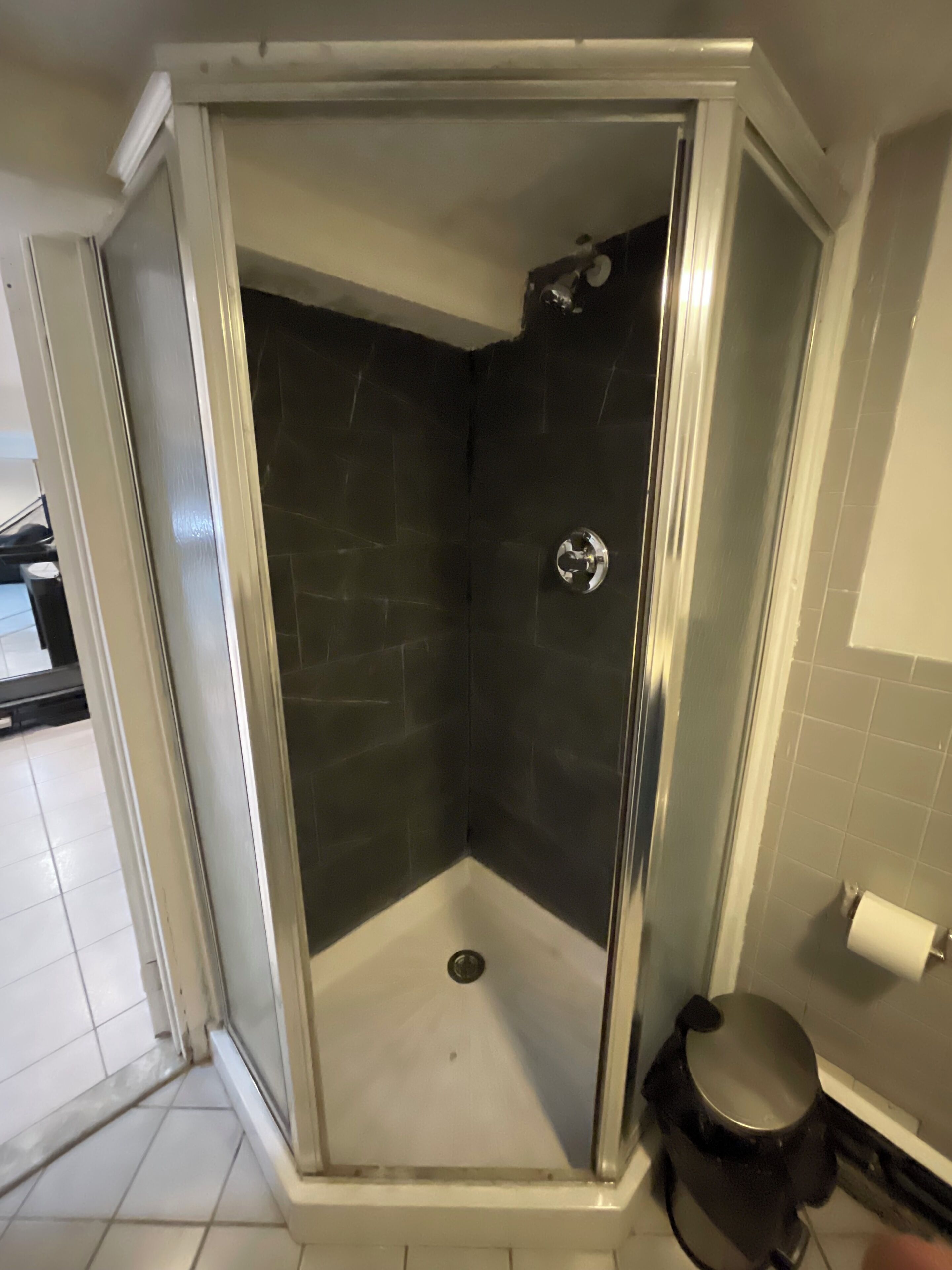 Une douche fermée par des parois de verre, avec des carreaux de couleur foncée, un sol blanc et des toilettes visibles sur la droite.