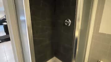 Une douche fermée par des parois de verre, avec des carreaux de couleur foncée, un sol blanc et des toilettes visibles sur la droite.
