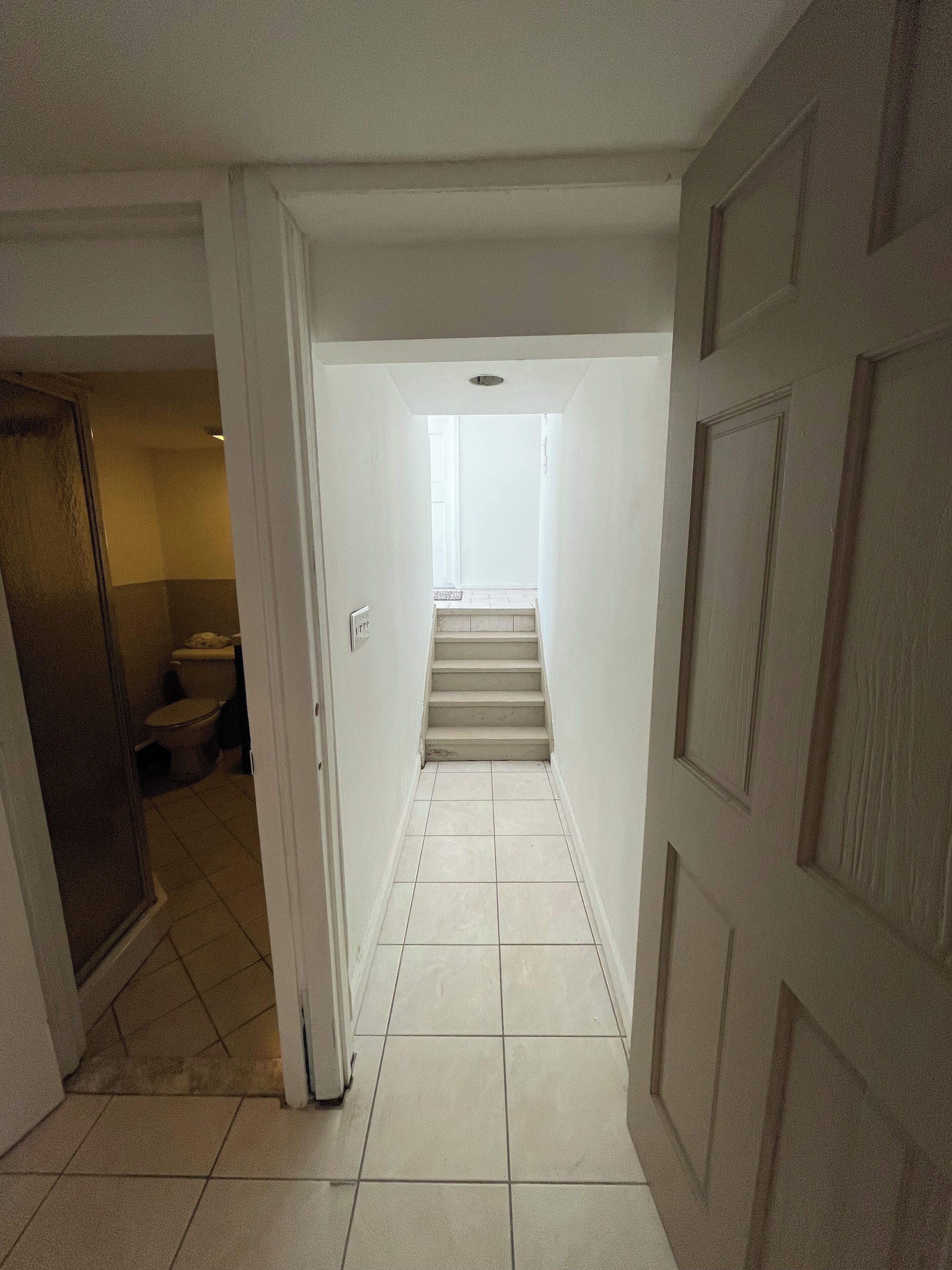 A narrow hallway with a staircase leading to an upper floor. The walls are white, and the floor is tiled.